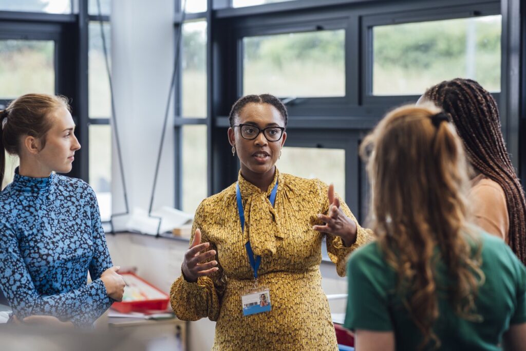 A shot of a mid-adult secondary school teacher talking with her sixth form students in class, they are wearing casual clothing and discussing what they learned in class