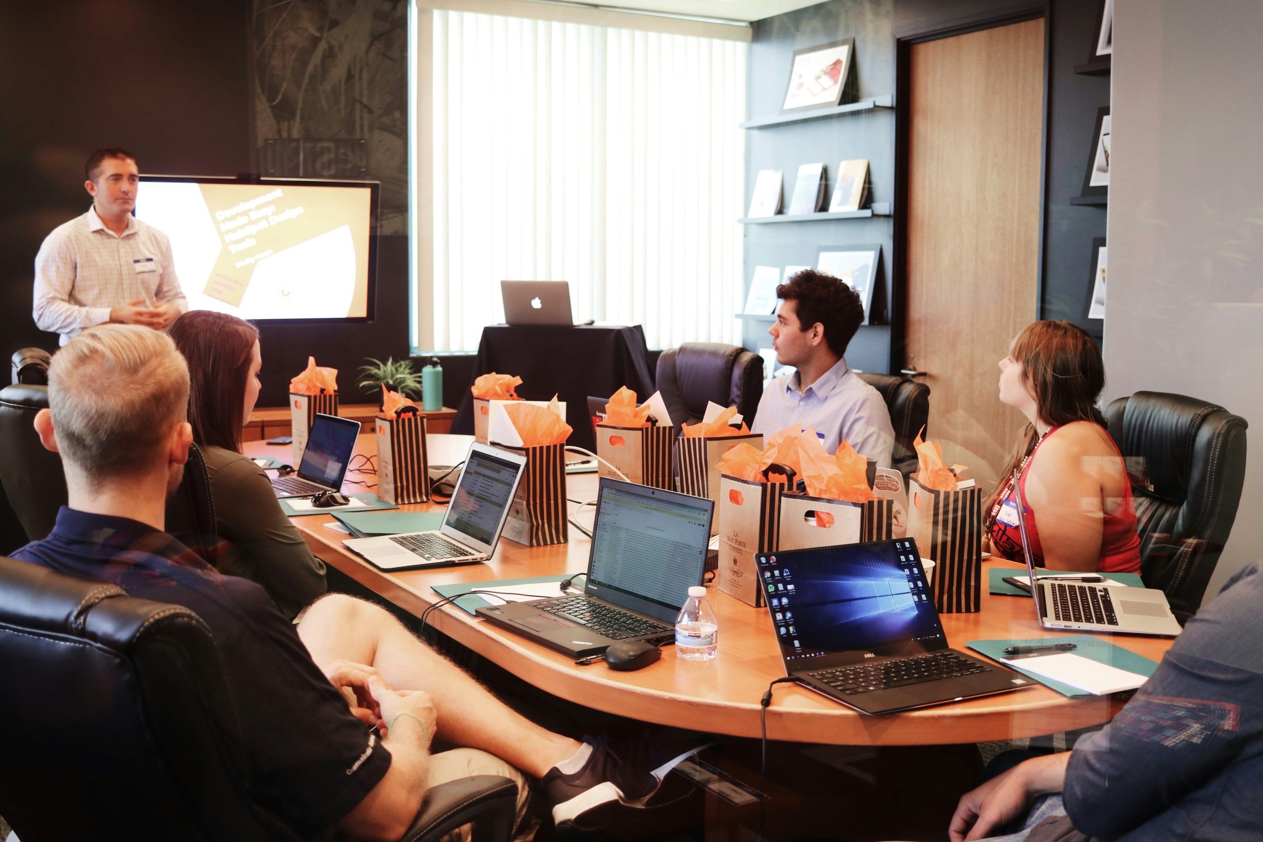 meeting room with people sat around large table at laptops watching person giving presentation