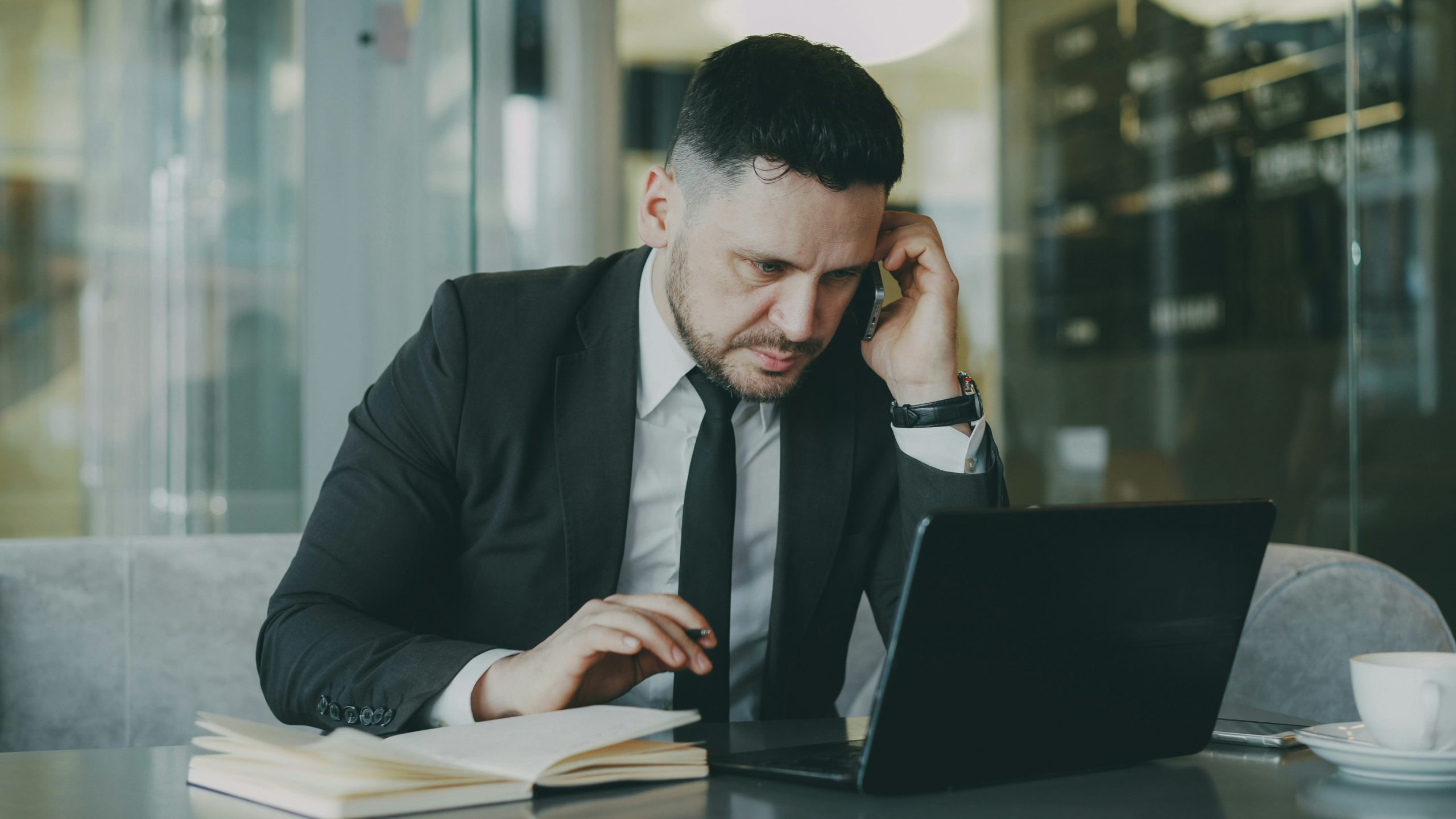 man at laptop on the phone looking stressed with notebook open beside him in an office