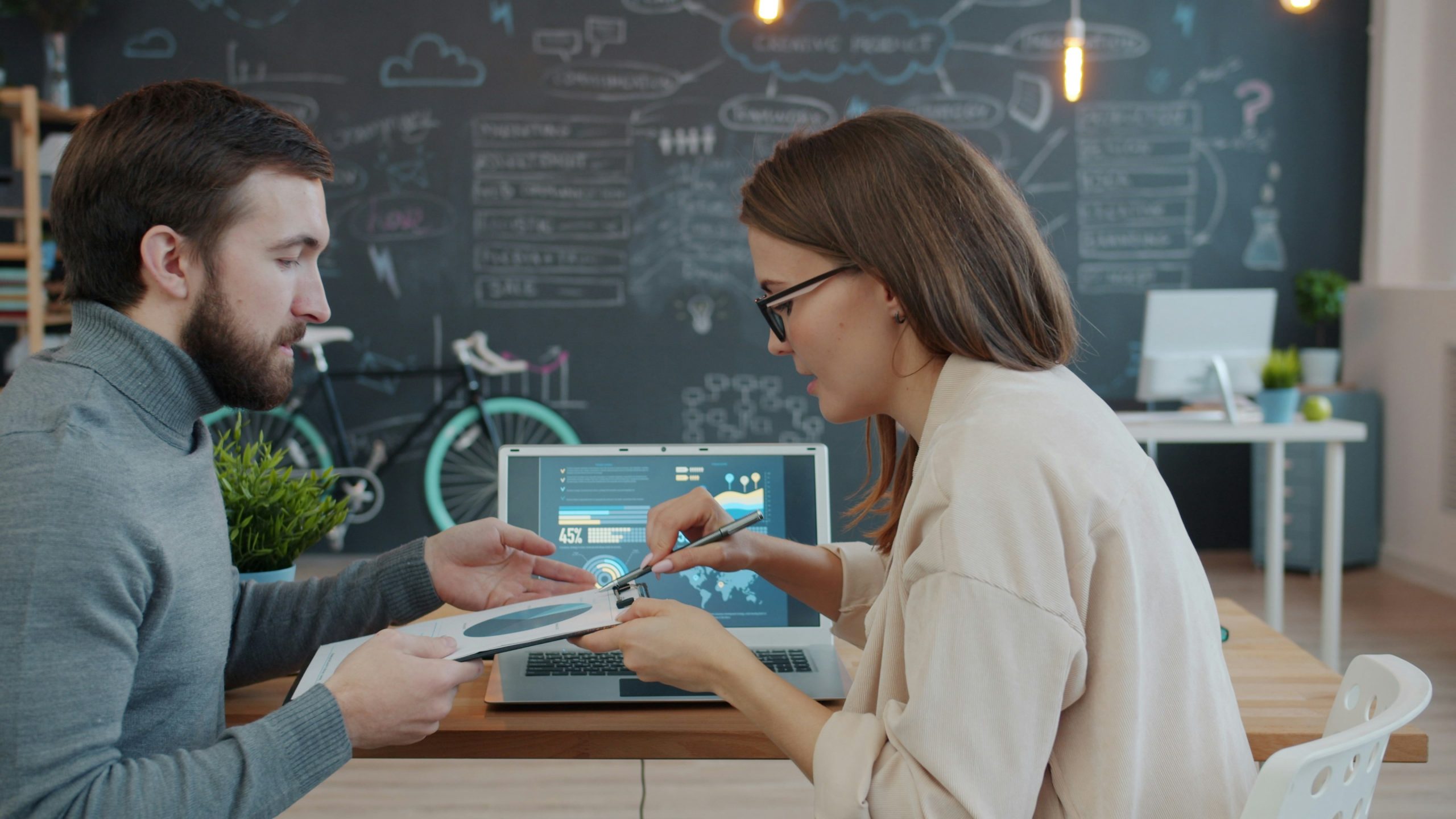 man and woman discussing business data with clipboard and laptop
